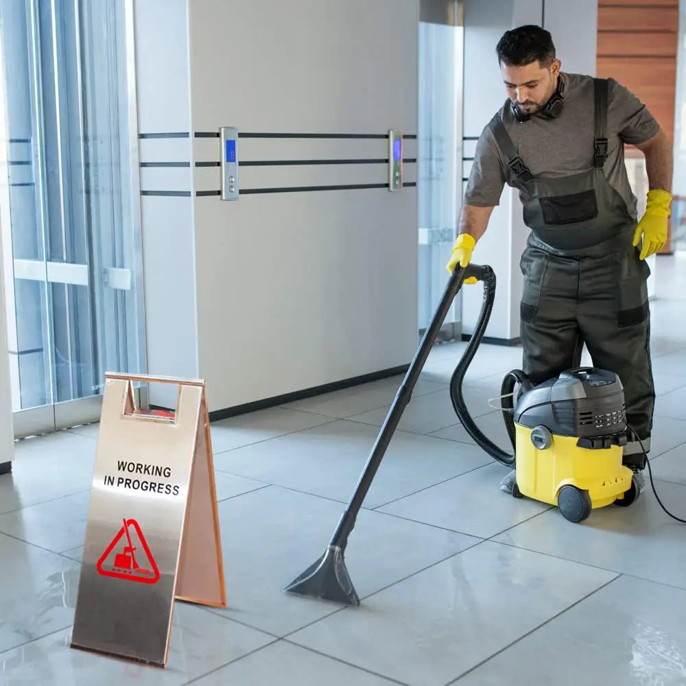 Person cleaning a floor with a vacuum cleaner, standing next to a 'Working In Progress' sign.